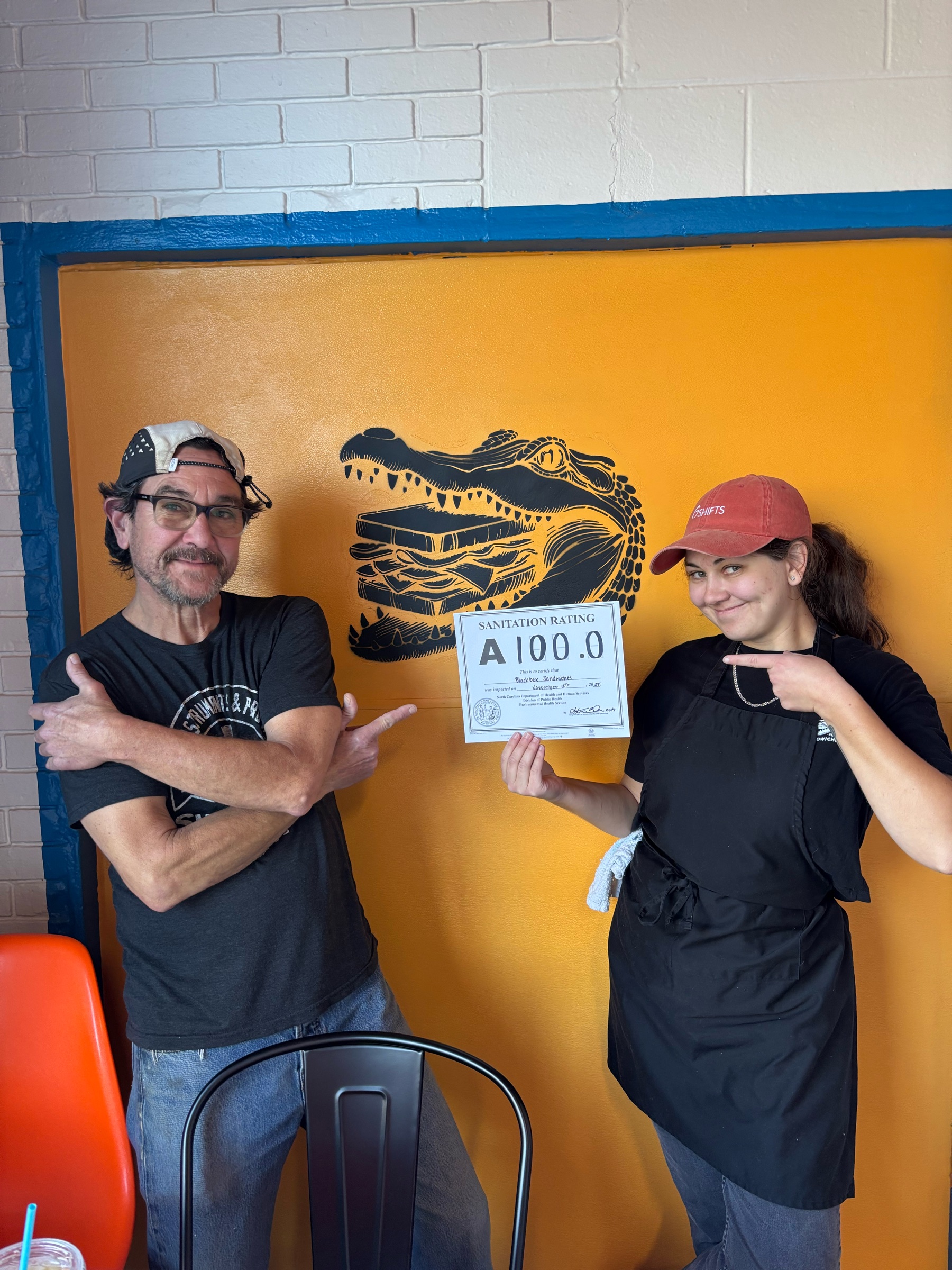 Claire and Shawn Mull holding up a 100.0 health inspection score in front of the alligator mural